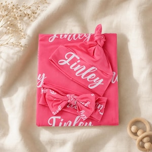 May include: A bright pink baby swaddle blanket, a knotted beanie, and a matching bow headband. Each item features the name Finley printed in a white cursive script repeatedly across the fabric. The set is displayed as a folded collection.