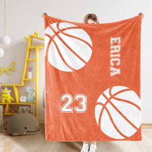 May include: An orange blanket with two large white basketballs, the name "ERICA" in white block letters, and the number "23". The blanket is made of a soft material, ideal for keeping warm.