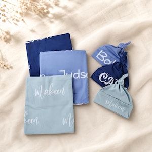 May include: Three folded rectangular blankets and three matching knotted baby hats in shades of blue. The items feature white printed names including Makeen, Judson, and others. The fabric is smooth and solid-colored, arranged in a layered composition.