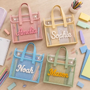 May include: Four square transparent mesh tote bags with two handles and snap closures. Each bag features a unique name in raised, textured script lettering: Amelia in pink, Sophie in white, Noah in white, and Mason in yellow. The bags contain stationery.