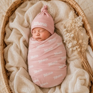 May include: A dusty rose baby swaddle blanket and matching knotted hat set, both featuring the name Athena printed in a white script font repeatedly across the fabric. The swaddle is wrapped snugly around a sleeping infant.