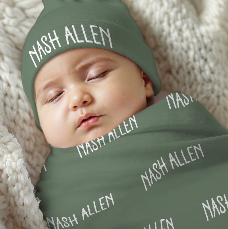May include: A sage green baby beanie and matching swaddle blanket set. Both items feature the name "NASH ALLEN" printed in a white, sans-serif font. The beanie has a folded brim, and the swaddle is wrapped around the infant's body.