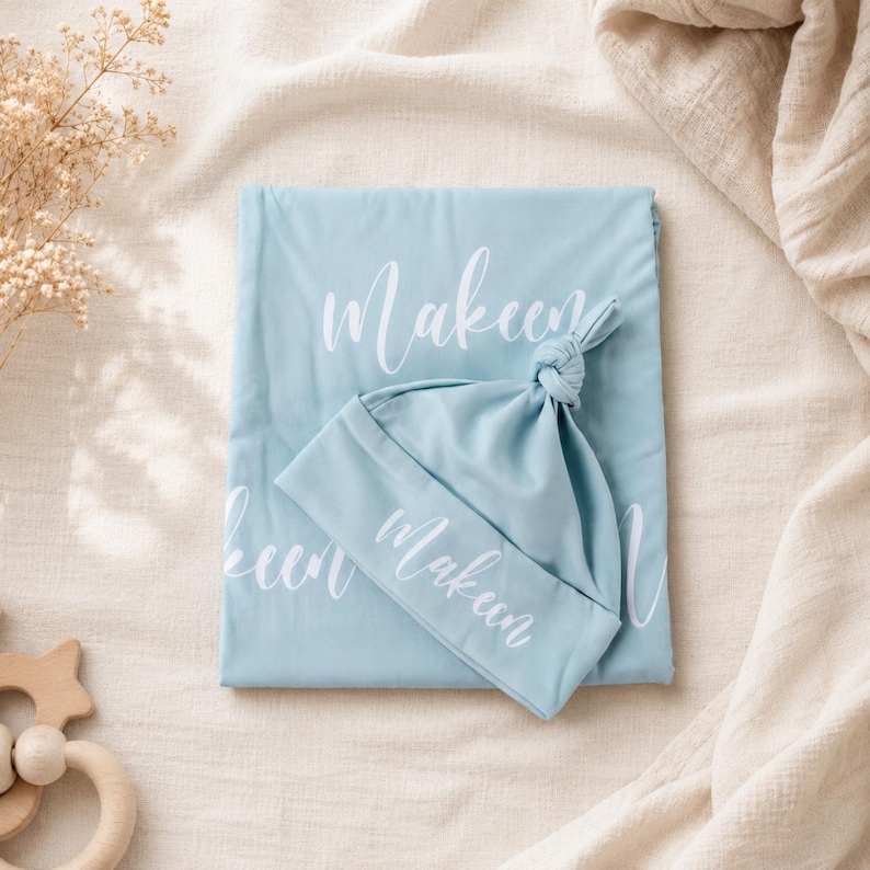 May include: A light blue baby blanket and a matching knotted hat, both featuring the name Makeen printed in white script. The fabric is smooth and solid in color, with the name repeated across the surface of the blanket and on the hat cuff.