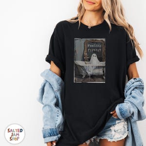 May include: Black t-shirt featuring a framed image of a ghost in a bathtub. The image has the text "Feeling filthy?" above the ghost. The shirt is part of the Salted Jam Shop collection.