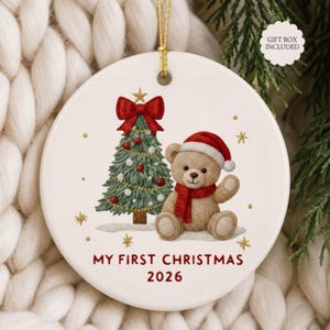 Può includere: Una decorazione rotonda in ceramica bianca con un cordino dorato. Il design raffigura un albero di Natale verde con un fiocco rosso, un orsacchiotto marrone con cappello e sciarpa rossi e la scritta "MY FIRST CHRISTMAS 2026" in rosso.