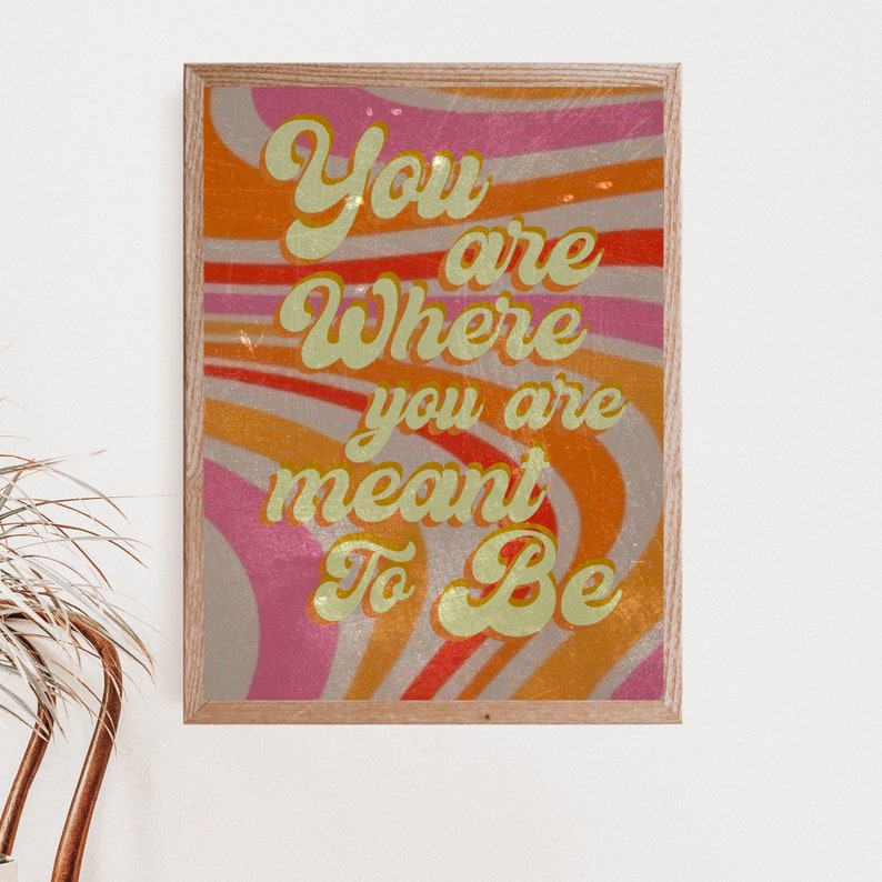 May include: A framed print with a retro-style design featuring a swirling pattern in shades of orange, pink, and white. The text "You are where you are meant to be" is written in a gold font.