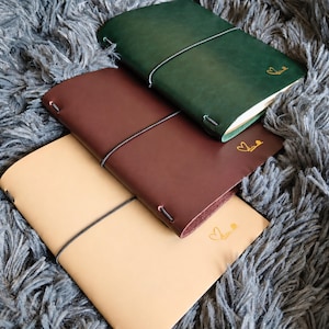 Peut inclure: Trois carnets en cuir de couleur vert foncé, marron foncé et beige clair. Chaque carnet est maintenu par un fin élastique noir et présente un petit motif doré en forme de cœur et de ligne embossé sur la couverture, près du coin inférieur droit.