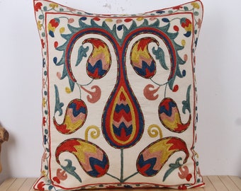 May include: A white throw pillow with a colorful embroidered design of stylized paisley shapes in shades of red, blue, yellow, and pink.