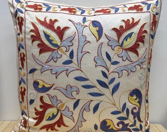 May include: Cream-colored square pillow with intricate floral embroidery in red, blue, and yellow. The design features stylized flowers and leaves, framed by a decorative border. A decorative accent for a sofa or bed.