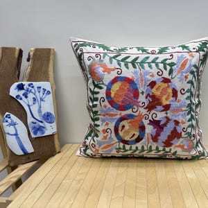 May include: White cotton pillow cover with embroidered floral design in shades of green, orange, pink, blue, and red. The design features four stylized flowers with intricate details.