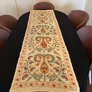 Könnte beinhalten: Ein weißer Tischläufer mit einem floralen Muster, das in Rot, Grün und Gelb gestickt ist. Der Läufer liegt auf einem schwarzen Tisch mit braunen Stühlen.