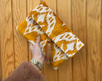 May include: A mustard yellow clutch with a white ikat pattern and a gold tassel. The clutch is made of velvet and has a flap closure.
