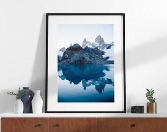 May include: A framed photograph of a mountain range reflected in a still body of water. The mountains are a dark blue and gray color, and the water is a light blue. The photograph is hanging on a wall above a wooden cabinet.