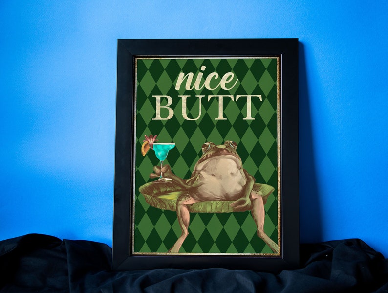May include: A framed print with a green background and a diamond pattern. The print features a cartoon frog sitting on a lily pad with a cocktail in its hand. The text "nice BUTT" is written in large letters above the frog.