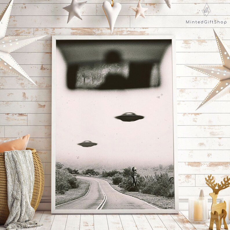 May include: A black and white photo of a road with two flying saucers in the sky. The photo is framed in a white frame and is hanging on a white wall.