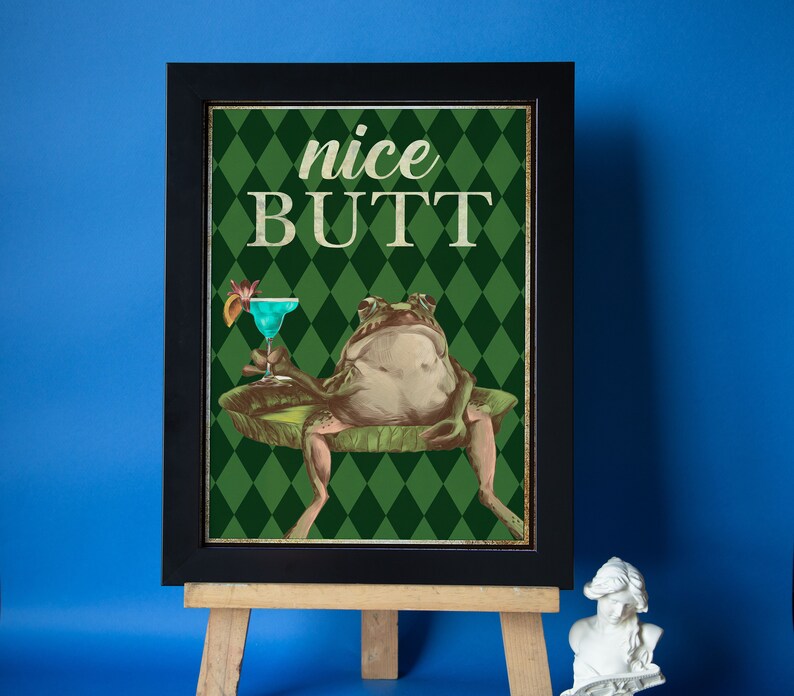 May include: A framed print with a green and white diamond pattern background. A cartoon frog sits on a lily pad holding a blue cocktail. The text "nice BUTT" is printed in white on the print.
