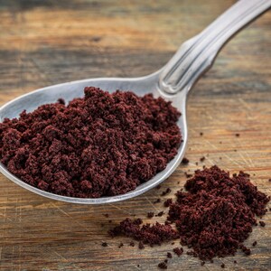 May include: A silver spoon filled with a heaping amount of dark red acai powder. A small amount of powder is scattered on the wooden surface.