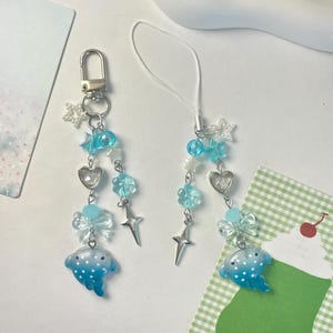 Op de afbeelding: Twee bedelarmbanden met een hemels thema. Elke bedel heeft een zilverkleurige sluiting of touwtje, heldere en blauwe kralen, ster- en hartbedels en een blauwe walvisvormige bedel. De bedels liggen op een witte achtergrond met decoratieve kaarten.