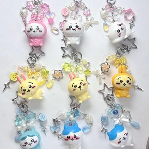 May include: Nine decorative keychains featuring cartoon characters in various colours including pink, white, yellow, and blue. Each keychain includes a silver star-shaped clasp, colourful beads, small charms shaped like bows, hearts, and stars.