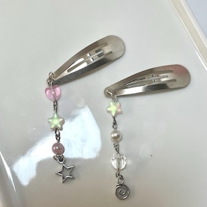 May include: Two silver hair clips with dangling charms. One clip has pink heart and star charms, the other has pearl, clear bead, and swirl charms. The charms are attached with silver chains.