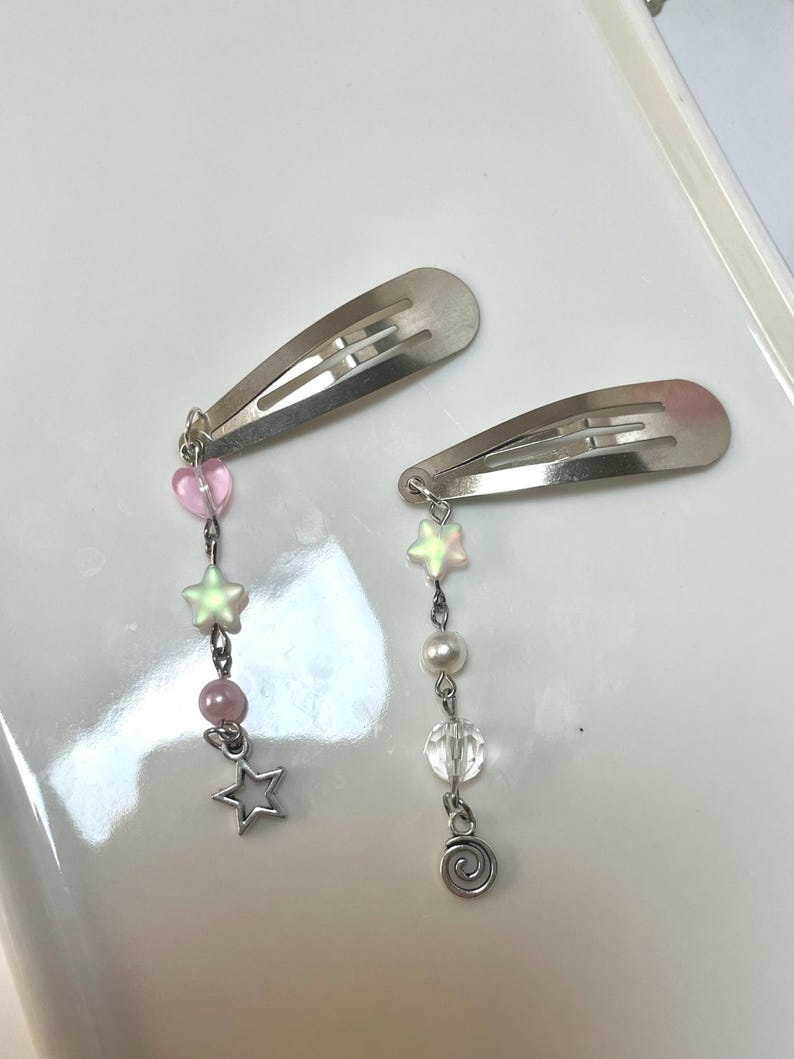 May include: Two silver hair clips with dangling charms. One clip has pink heart and star charms, the other has pearl, clear bead, and swirl charms. The charms are attached with silver chains.