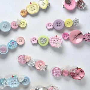 May include: A collection of hair clips featuring various colorful buttons in shapes like hearts, apples, and flowers. The buttons are arranged in rows on metal clips, showcasing patterns including gingham, solid colors, and glittery accents in pink, blue, yellow, and green.