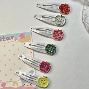 Pode incluir: Seis grampos de cabelo prateados, cada um adornado com um enfeite colorido em forma de maçã. As maçãs apresentam vários padrões e cores, incluindo xadrez vermelho, xadrez verde e amarelo com bolinhas brancas. Um cartão decorativo com estrelas e a palavra "star" é visível.