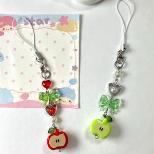 May include: Two decorative phone charms with white cords. One features a red apple, green bow, and red heart charms. The other has a green apple, green bow, and a heart charm. Both have silver-tone hardware.