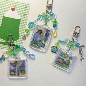 Könnte beinhalten: Drei quadratische Schlüsselanhänger aus klarem Acryl mit Collage-Einlagen in Blau- und Grüntönen, darunter Computer-Icons und Naturszenen. Jeder Anhänger ist an einem silbernen Karabinerhaken befestigt und mit verschiedenen blauen, grünen und transparenten Perlen in Stern-, Blumen- und Schmetterlingsform verziert.