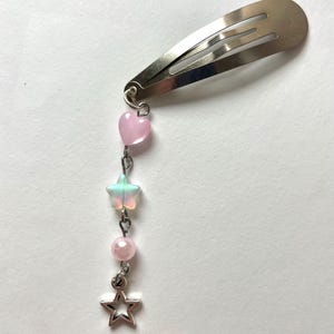 May include: Silver metal hair clip with a dangling charm. The charm includes a pink heart, a translucent star, a pink bead, and a silver star-shaped charm. The clip is designed to secure hair.