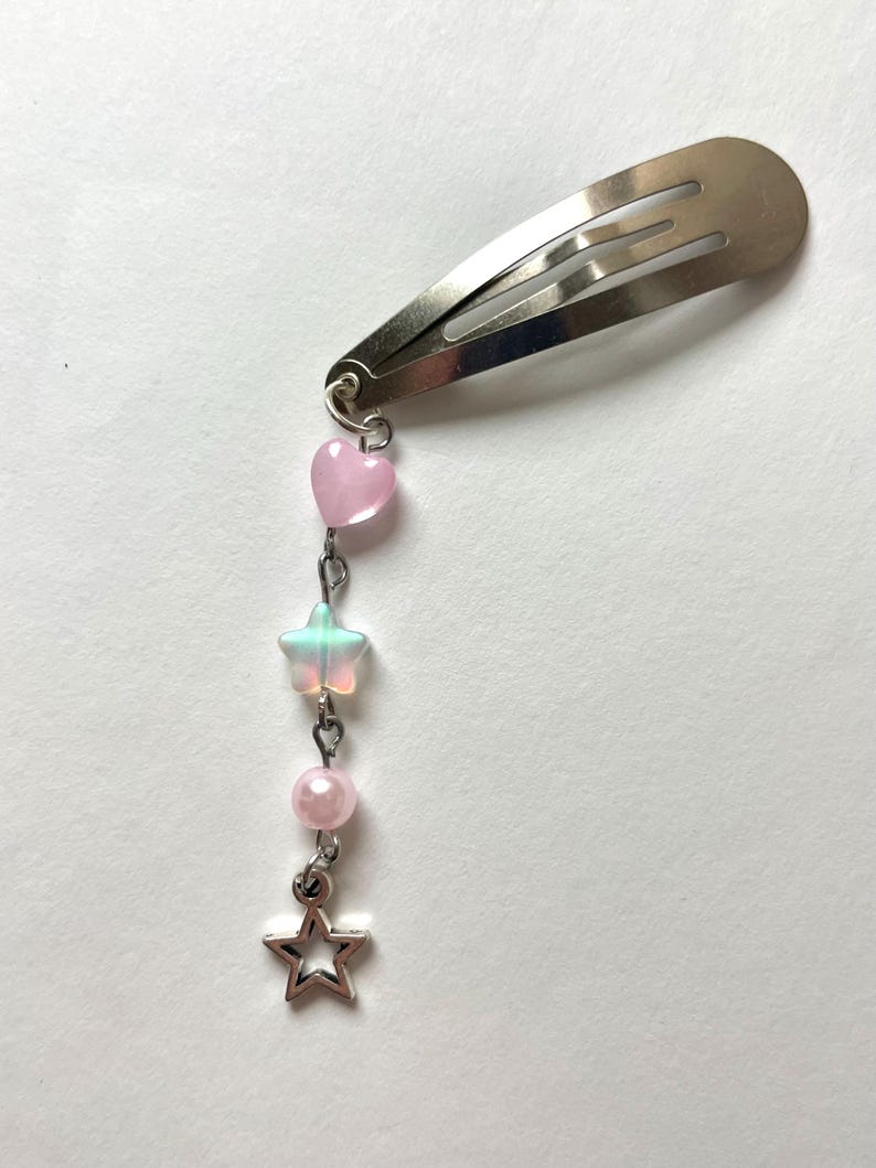 May include: Silver metal hair clip with a dangling charm. The charm includes a pink heart, a translucent star, a pink bead, and a silver star-shaped charm. The clip is designed to secure hair.