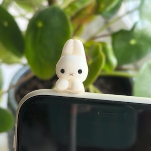 May include: A small, white, glossy ceramic figurine of a rabbit with long ears, two black circular eyes, and a small cross-shaped mouth. The rabbit figurine is attached to the top edge of a smartphone with a black screen and a light-colored frame.