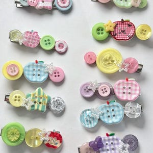 Pode incluir: Uma coleção de dez presilhas de cabelo com botões variados, pingentes em forma de estrela e peças em formato de maçã em cores como rosa, verde, azul, amarelo e roxo. Cada presilha apresenta um arranjo único de botões e pequenos detalhes de estrela.