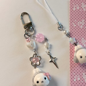 K&ouml;nnte beinhalten: Ein Handy-Anh&auml;nger mit einer wei&szlig;en Hasenkopf-Figur, die graue Kopfh&ouml;rer und eine rosa Schleife tr&auml;gt. Der Anh&auml;nger besteht aus einem silbernen Karabinerverschluss, einer wei&szlig;en Kordel und verschiedenen Perlen, darunter eine rosa Blume, ein wei&szlig;es Herz und ein silberner Stern.