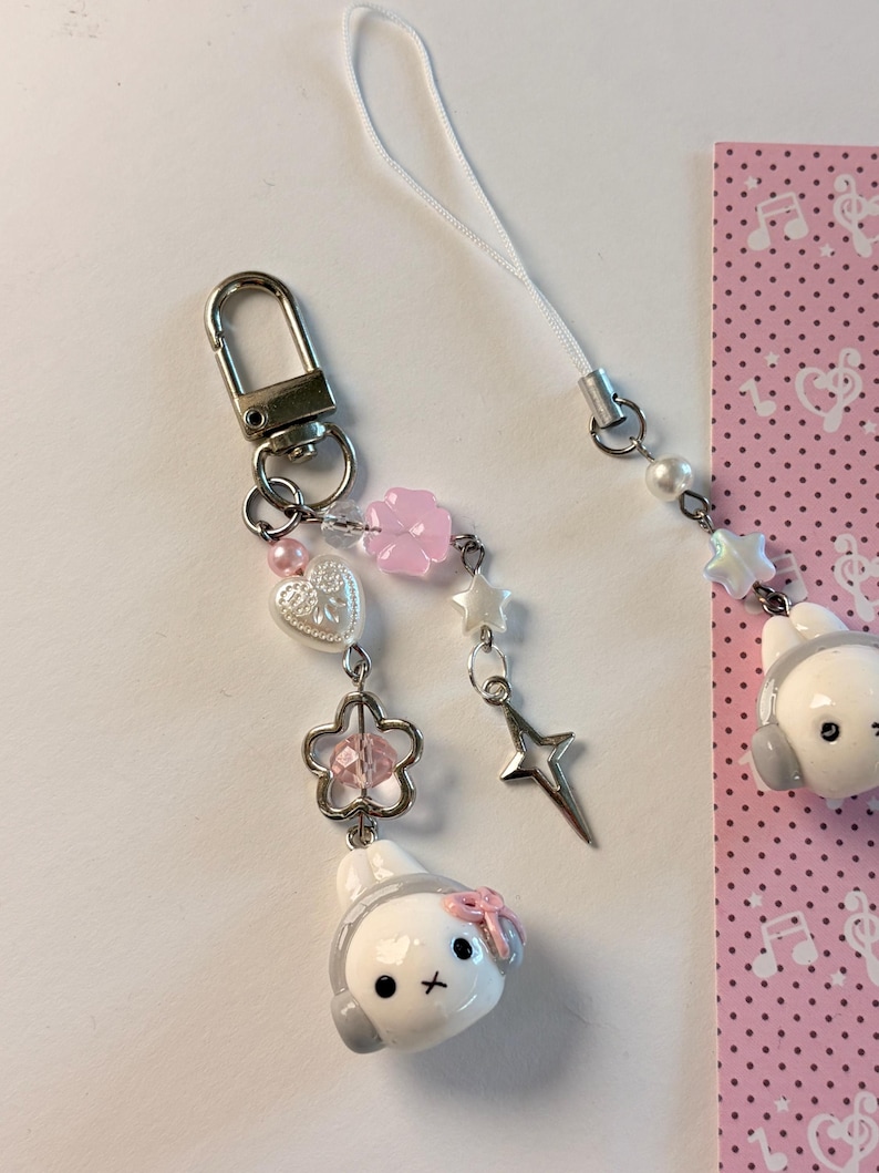 K&ouml;nnte beinhalten: Ein Handy-Anh&auml;nger mit einer wei&szlig;en Hasenkopf-Figur, die graue Kopfh&ouml;rer und eine rosa Schleife tr&auml;gt. Der Anh&auml;nger besteht aus einem silbernen Karabinerverschluss, einer wei&szlig;en Kordel und verschiedenen Perlen, darunter eine rosa Blume, ein wei&szlig;es Herz und ein silberner Stern.