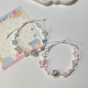 Op de afbeelding: Twee delicate armbanden met witte koorden, versierd met kralen en bedels. Eén heeft vlinder- en hartbedels, de andere heeft ster- en hartbedels. De armbanden liggen op een wit oppervlak naast een decoratieve kaart.