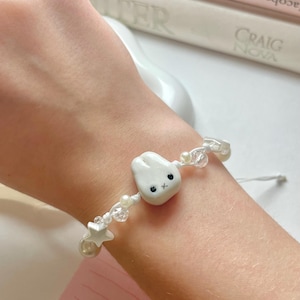 May include: A white beaded bracelet with a cartoon tooth charm with black eyes and a small star charm. The bracelet is strung on white cord and includes clear and pearl-like beads.