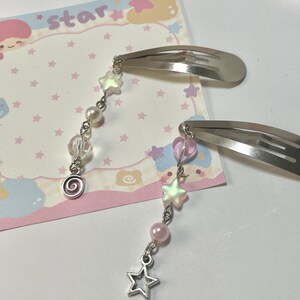 May include: Two silver hair clips with dangling charms. Each charm features a star, beads, and a spiral design. The background includes a pink and white card with the word "star" and star illustrations.
