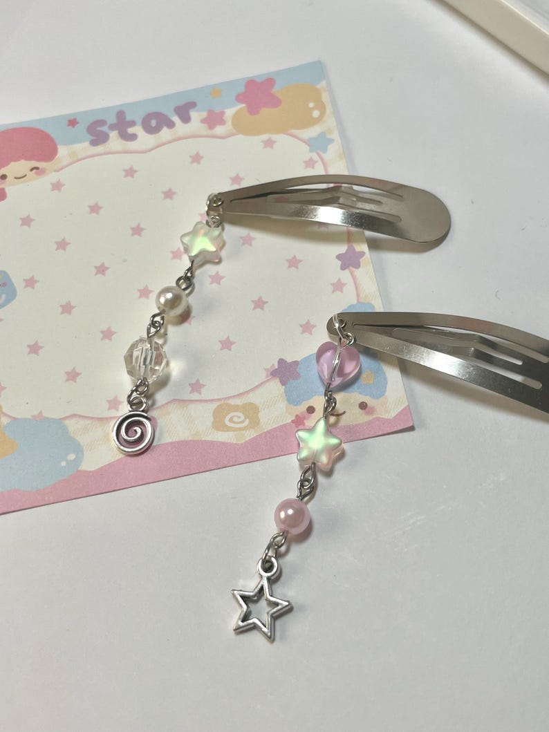 May include: Two silver hair clips with dangling charms. Each charm features a star, beads, and a spiral design. The background includes a pink and white card with the word "star" and star illustrations.