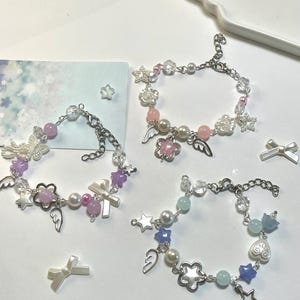 May include: Three charm bracelets with star, flower, and angel wing charms. The bracelets feature pearls and pink, purple, and blue beads. Silver chains and charms complete the design. The bracelets are suitable for wrists of various sizes.