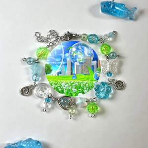 Könnte beinhalten: Ein Armband mit einem zentralen kreisförmigen Anhänger mit einer Stadtlandschaft und der Erde. Das Armband enthält blaue, grüne und klare Perlen sowie Anhänger in Form eines Schmetterlings, einer Blume und eines Herzens. Zwei blaue Wal-Anhänger sind ebenfalls vorhanden.
