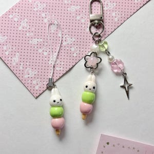 May include: Two charms featuring a white rabbit head atop a stack of green and pink spheres. One charm has a white cord loop, while the other includes a silver metal lobster clasp, a clear star bead, a pink flower bead, and a silver star charm.