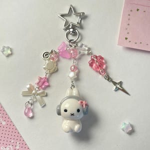 Può includere: Un portachiavi stravagante con una figura di coniglietto bianco con cuffie e un fiocco rosa. Il portachiavi include un fermaglio a forma di stella, perline rosa e bianche, fiocchi e ciondoli a stella. Il design è pastello e carino.