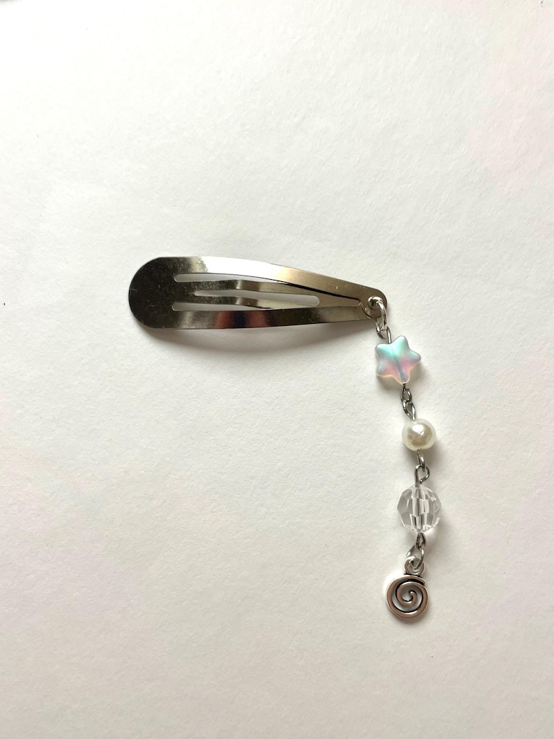May include: A silver hair clip with a dangling charm. The charm includes a pearlescent star, a white pearl, a clear faceted bead, and a silver spiral. The clip is designed to secure hair.