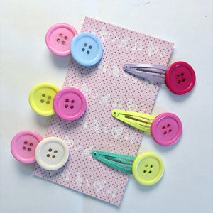 Pode incluir: Uma coleção de ganchos de cabelo coloridos com botões redondos de quatro furos em tons de rosa, amarelo, azul, branco e verde, fixados em clipes de metal, apresentados num cartão retangular com fundo rosa e notas musicais brancas.