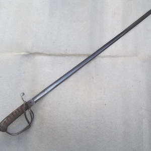 Pode incluir: Uma espada de aço longa e fina com uma lâmina metálica escura e ponta afiada. O punho inclui uma empunhadura castanha canelada e um protetor de mão de metal curvo que se estende do pomo até à guarda, proporcionando proteção para a mão.