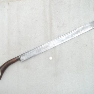 Puede incluir: Un machete largo y plateado con un mango curvo de madera. La hoja es rectangular con una punta redondeada. El mango tiene un diseño curvo con dos púas en el extremo. El machete está sobre una superficie clara.
