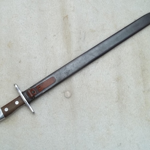 Op de afbeelding: Een vintage bajonet met een donker metalen lemmet en een houten handvat. Het handvat is vastgezet met een metalen bescherming en een leren riem. De bajonet zit in een metalen schede. Het kleurenschema is donkergrijs, bruin en zilver.