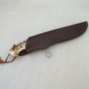 Puede incluir: Un cuchillo de hoja fija con mango de asta texturizado y una funda de cuero marrón con costuras decorativas. Un cordón fino de cuero marrón está unido al pomo. Una pequeña moneda de plata está colocada junto a la funda como referencia.