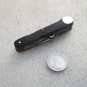 May include: A closed folding pocket knife with a black textured handle featuring a diamond pattern and a silver circular end cap, positioned next to a silver coin showing a profile of Queen Elizabeth II for scale.